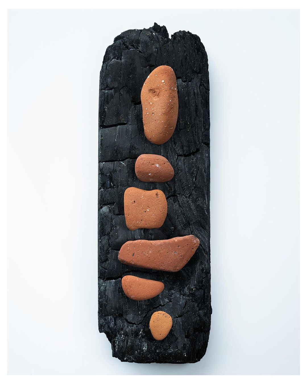 Arrangement of red stones balanced atop a dark, charred piece of wood against a plain white background in an abstract composition.