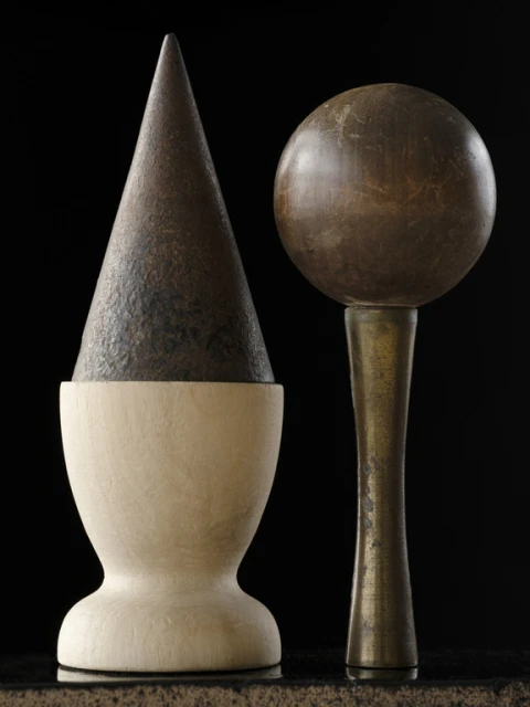 Still life featuring geometric wood sculptures with a cone on a pedestal and a sphere on a brass stem, set against a stark black background.