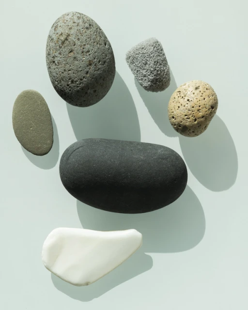 A collection of grey, black, white, and tan smooth rocks and stones with varying shapes and sizes arranged on a light blue surface.