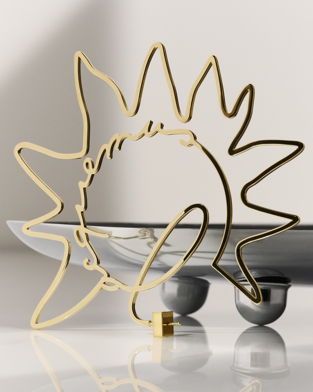 Gold wire art piece featuring a human face and sun shape, sitting atop a reflective white surface with a blurred white background.