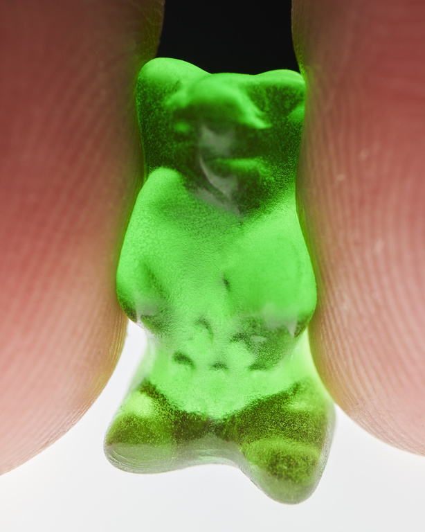 Close-up of a green gummy bear candy held between two fingers against a white background, detail of candy texture and translucence.