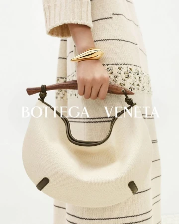 Close-up of a Bottega Veneta hobo bag with a wooden handle and a gold bracelet, held by an adult female model, against a neutral background.