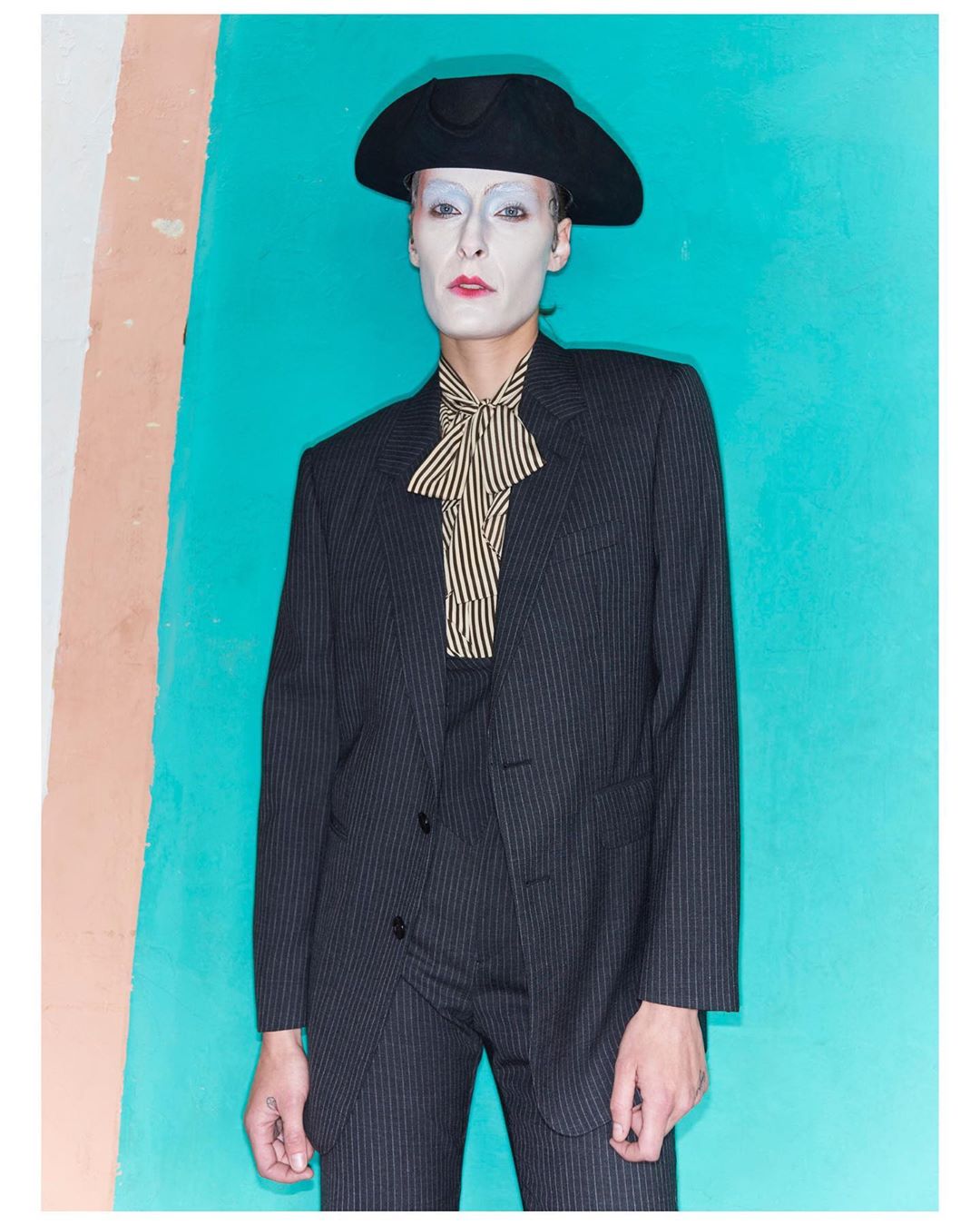 Portrait of a person in a black pinstripe suit and black hat, with white makeup, standing against a blue and peach-colored wall.