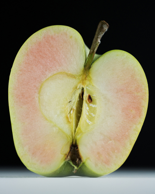 Studio shot of a halved green apple with pinkish flesh and stem against a dark background, showcasing its texture and form.