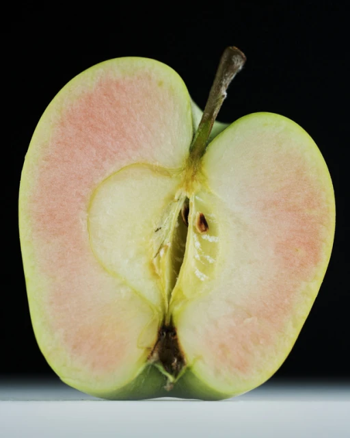 Studio shot of a halved green apple with pinkish flesh and stem against a dark background, showcasing its texture and form.