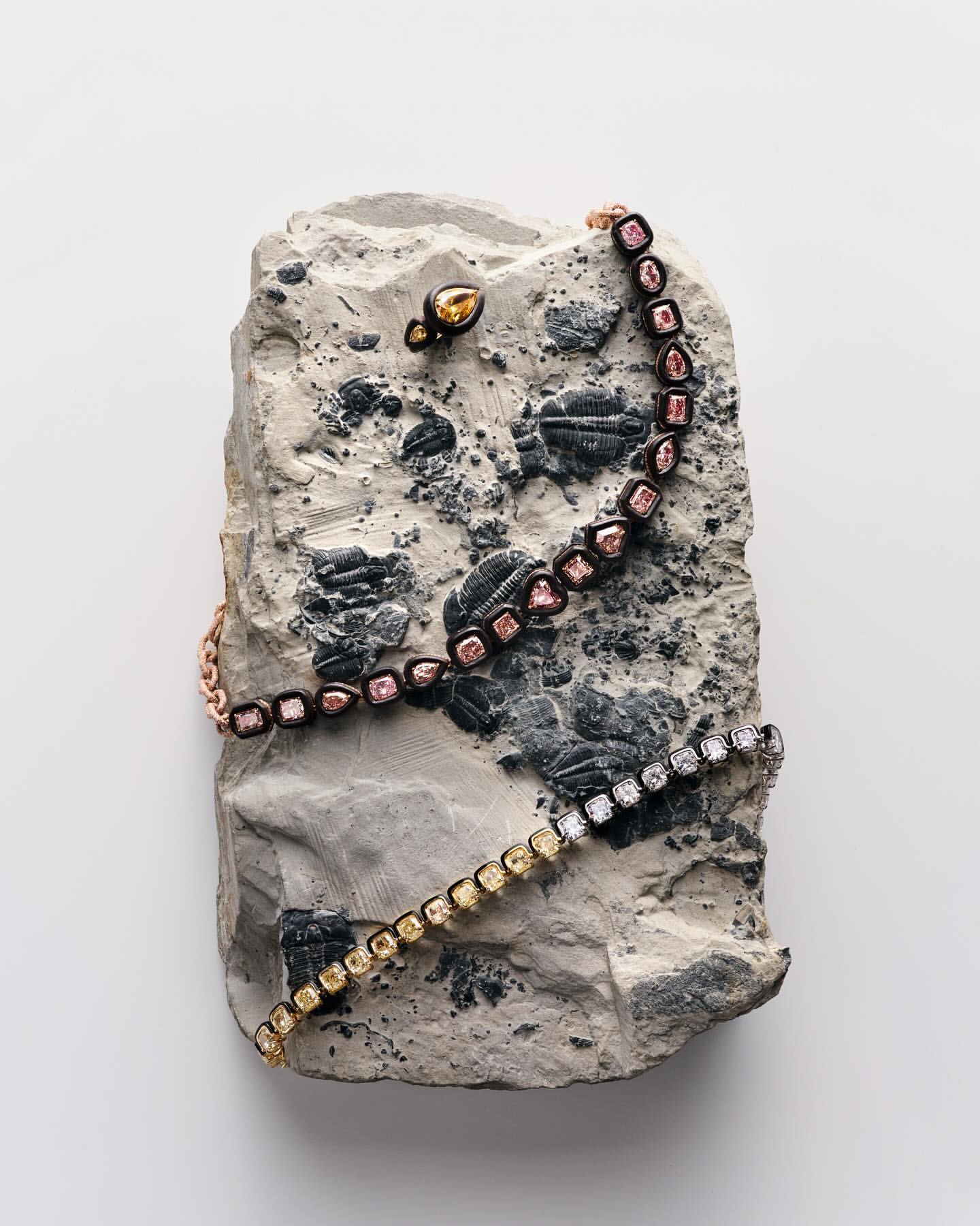 Arrangement of bracelets and an earring, featuring yellow and pink diamonds, artfully displayed on a textured gray stone against a white backdrop.