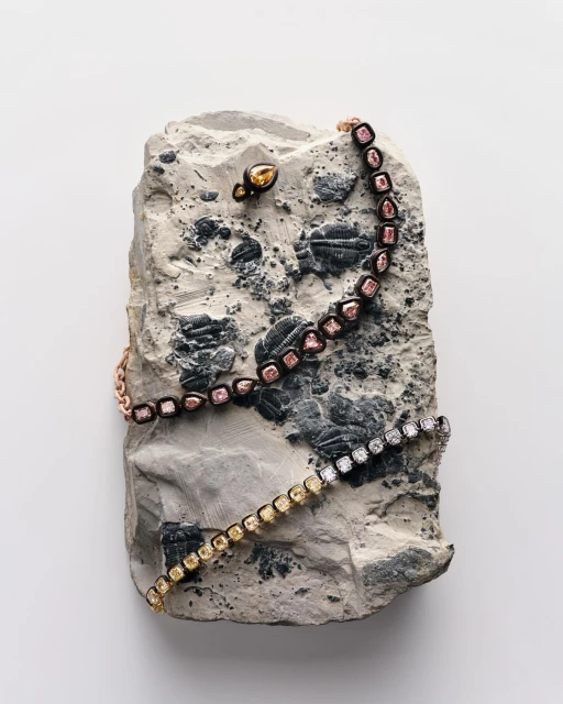 Arrangement of bracelets and an earring, featuring yellow and pink diamonds, artfully displayed on a textured gray stone against a white backdrop.