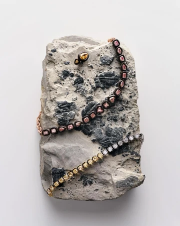 Arrangement of bracelets and an earring, featuring yellow and pink diamonds, artfully displayed on a textured gray stone against a white backdrop.