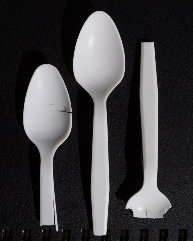 Studio shot of three damaged white plastic spoons of different sizes against a black background, showcasing wear and breakage.
