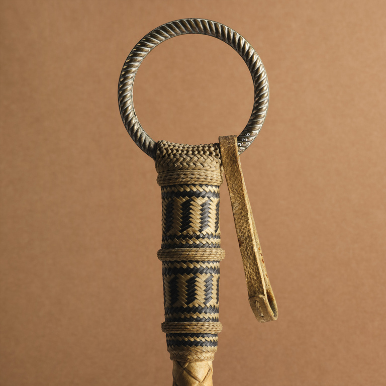 Close-up of a woven riding crop or polo whip with a metal ring, showcasing its detailed craftsmanship and design against a plain background.