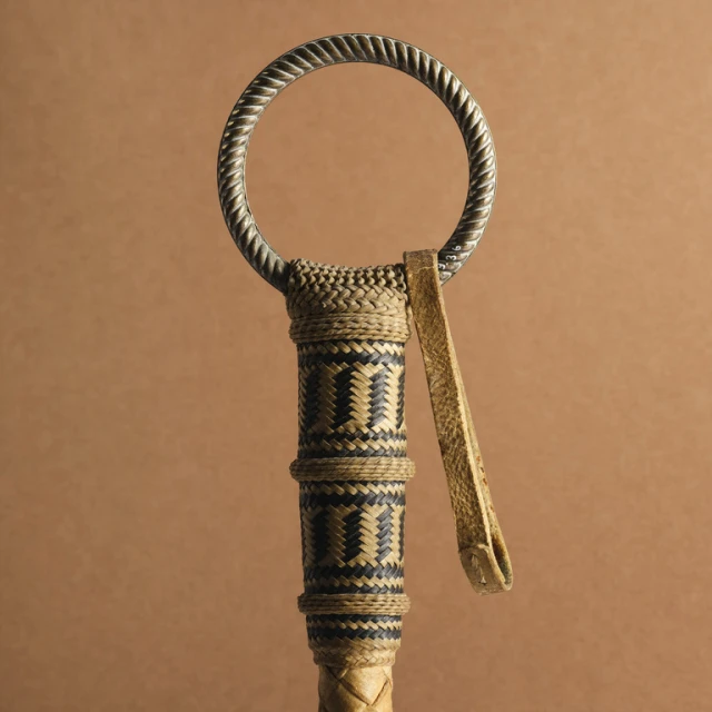 Close-up of a woven riding crop or polo whip with a metal ring, showcasing its detailed craftsmanship and design against a plain background.