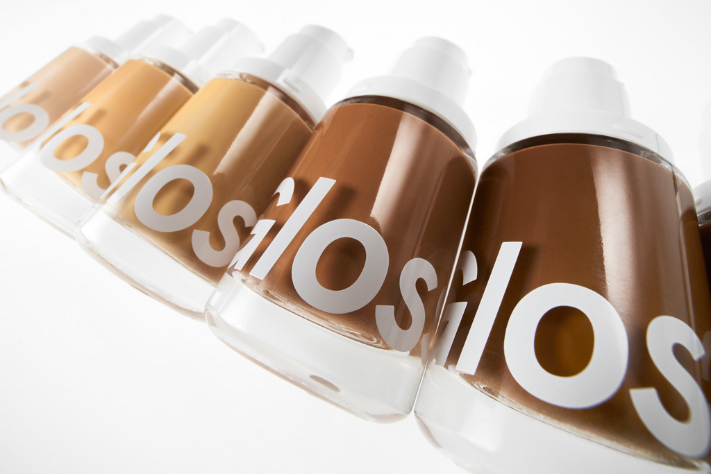 A row of glass cosmetic bottles with white caps, filled with different shades of liquid foundation against a white background.