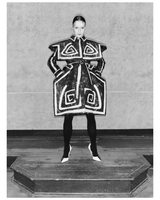 Monochrome portrait of a fashion model in a geometrically patterned outfit standing on a platform in front of a textured wall.