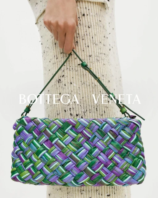 Close-up of a woven Bottega Veneta handbag in shades of green and purple, held by a person wearing a speckled, cream-colored sweater.