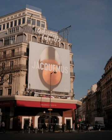 Exterior of Galeries Lafayette in Paris featuring a large Jacquemus advertisement, with architectural details and street scene under a blue sky.