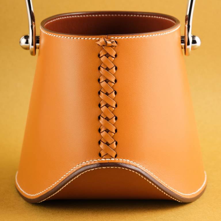 Close-up of a tan-colored leather bucket bag with silver hardware and braided leather detail against a golden-yellow background.