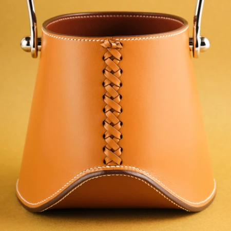 Close-up of a tan-colored leather bucket bag with silver hardware and braided leather detail against a golden-yellow background.