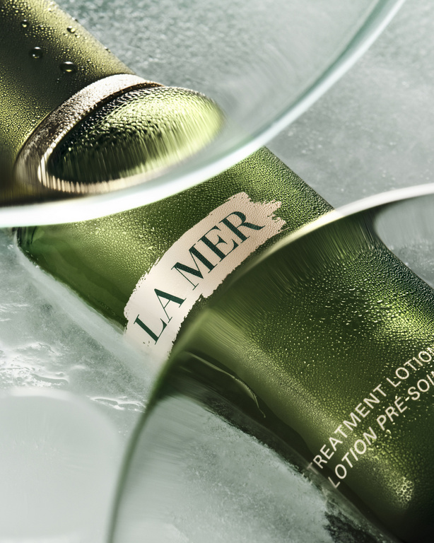 La Mer lotion product shot: A close-up of a green bottle with water droplets and the La Mer logo, reflecting in glass.