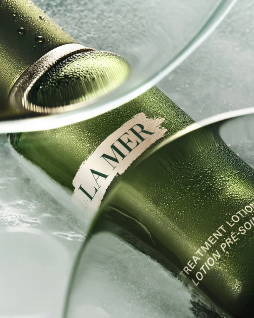 La Mer lotion product shot: A close-up of a green bottle with water droplets and the La Mer logo, reflecting in glass.