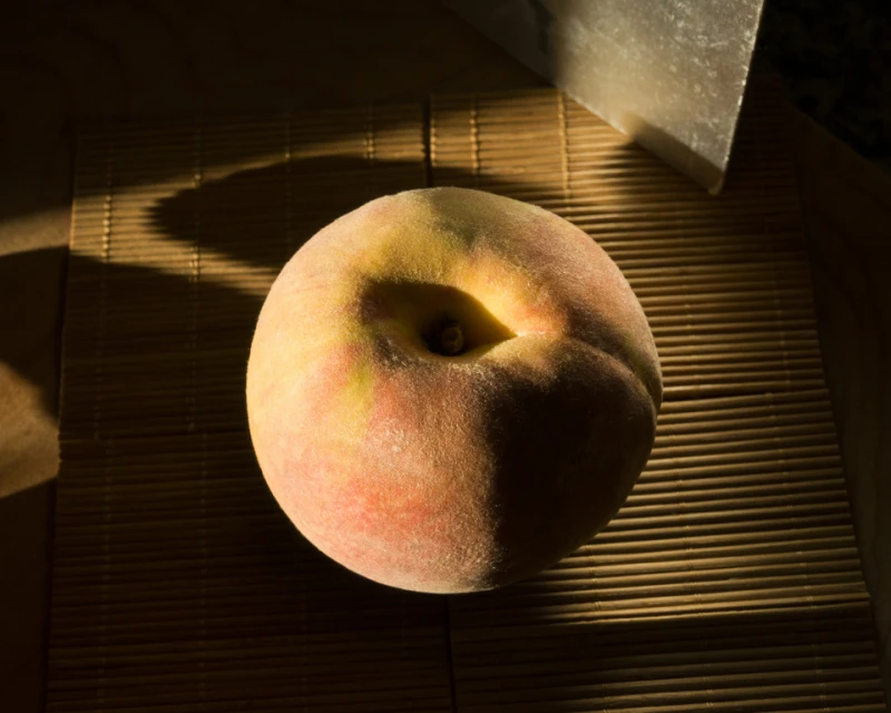 Still life studio shot of a ripe peach on a bamboo mat with a metallic object, showcasing the fruit's texture and warm colors.