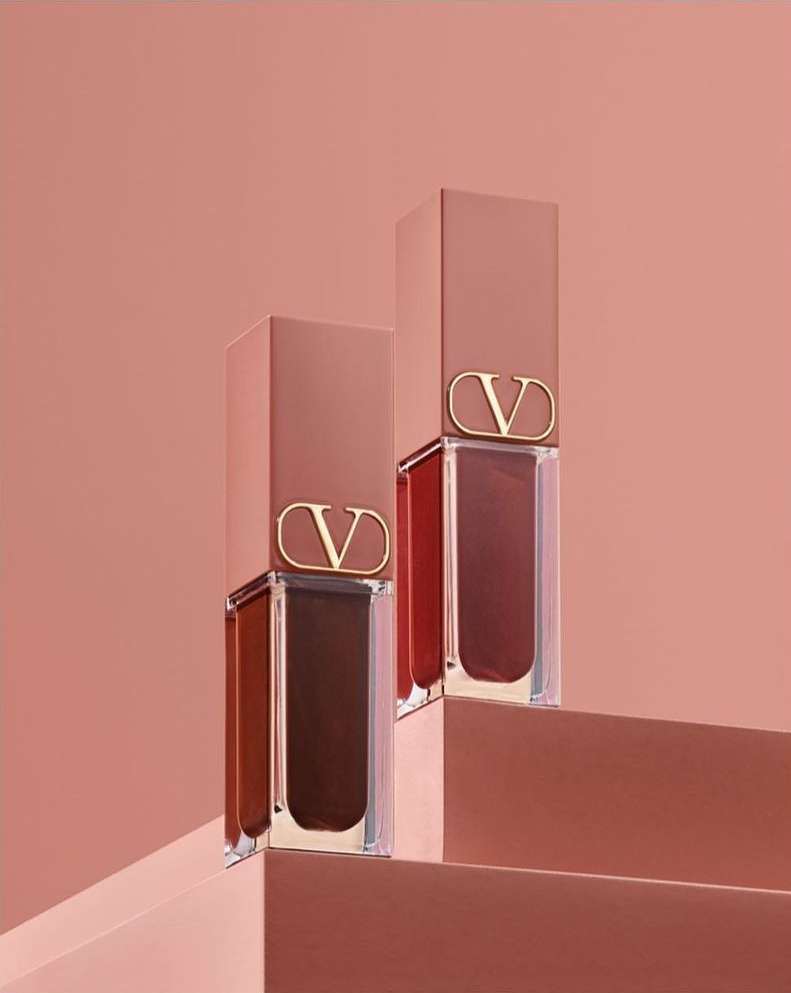 Two Valentino lipsticks in clear tubes and tan caps with a 'V' logo, displayed on tan steps against a matching monochromatic backdrop.