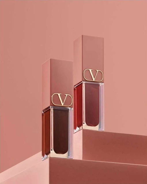 Two Valentino lipsticks in clear tubes and tan caps with a 'V' logo, displayed on tan steps against a matching monochromatic backdrop.