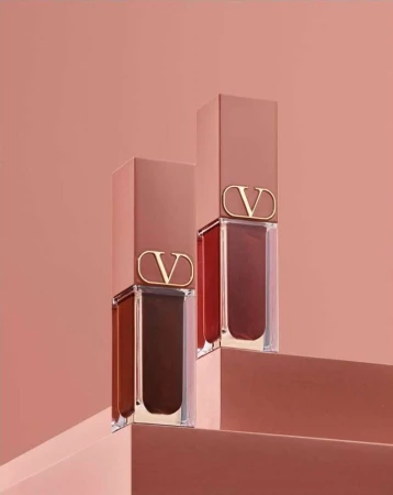 Two Valentino lipsticks in clear tubes and tan caps with a 'V' logo, displayed on tan steps against a matching monochromatic backdrop.