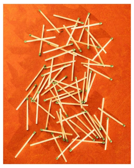 Scattered matches with green tips on orange surface create a textured pattern; minimalist studio shot with warm, inviting tones.