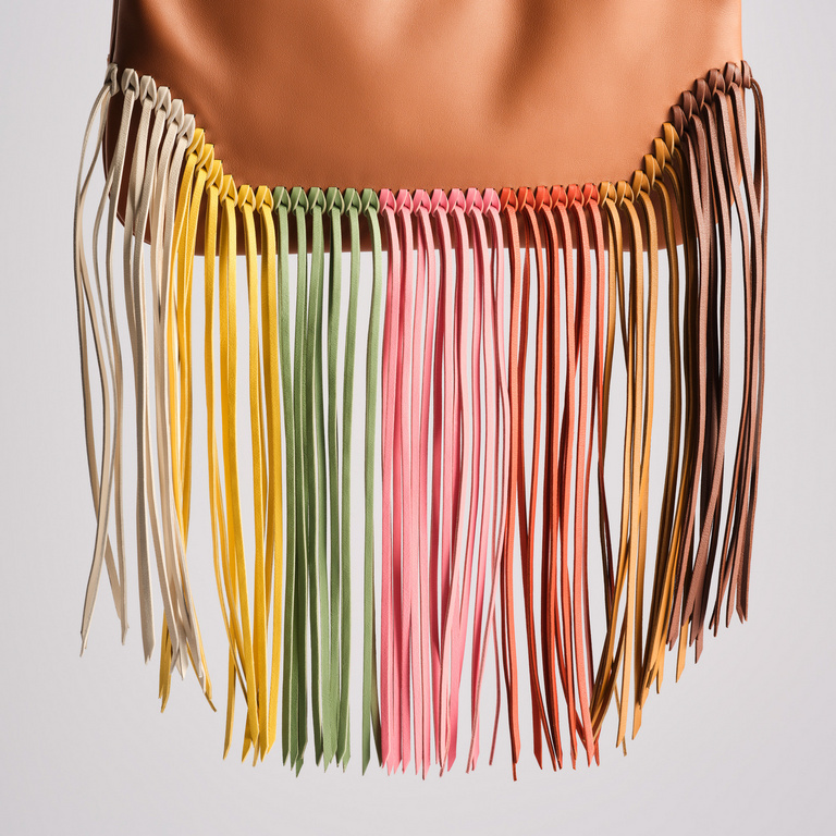 Close-up of a fringe textile or fiber art with vertical strands in beige, yellow, green, pink, orange, and brown against a white background.