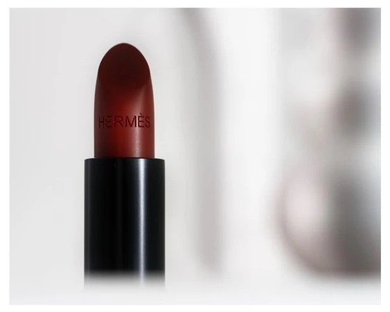 Close-up studio shot of a dark red Hermes lipstick tube against a soft, blurred white background. Beauty and fashion concept.