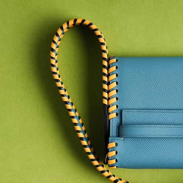 Close-up of a turquoise-colored leather handbag with a yellow and black braided handle on a green background, showcasing a luxury accessory.