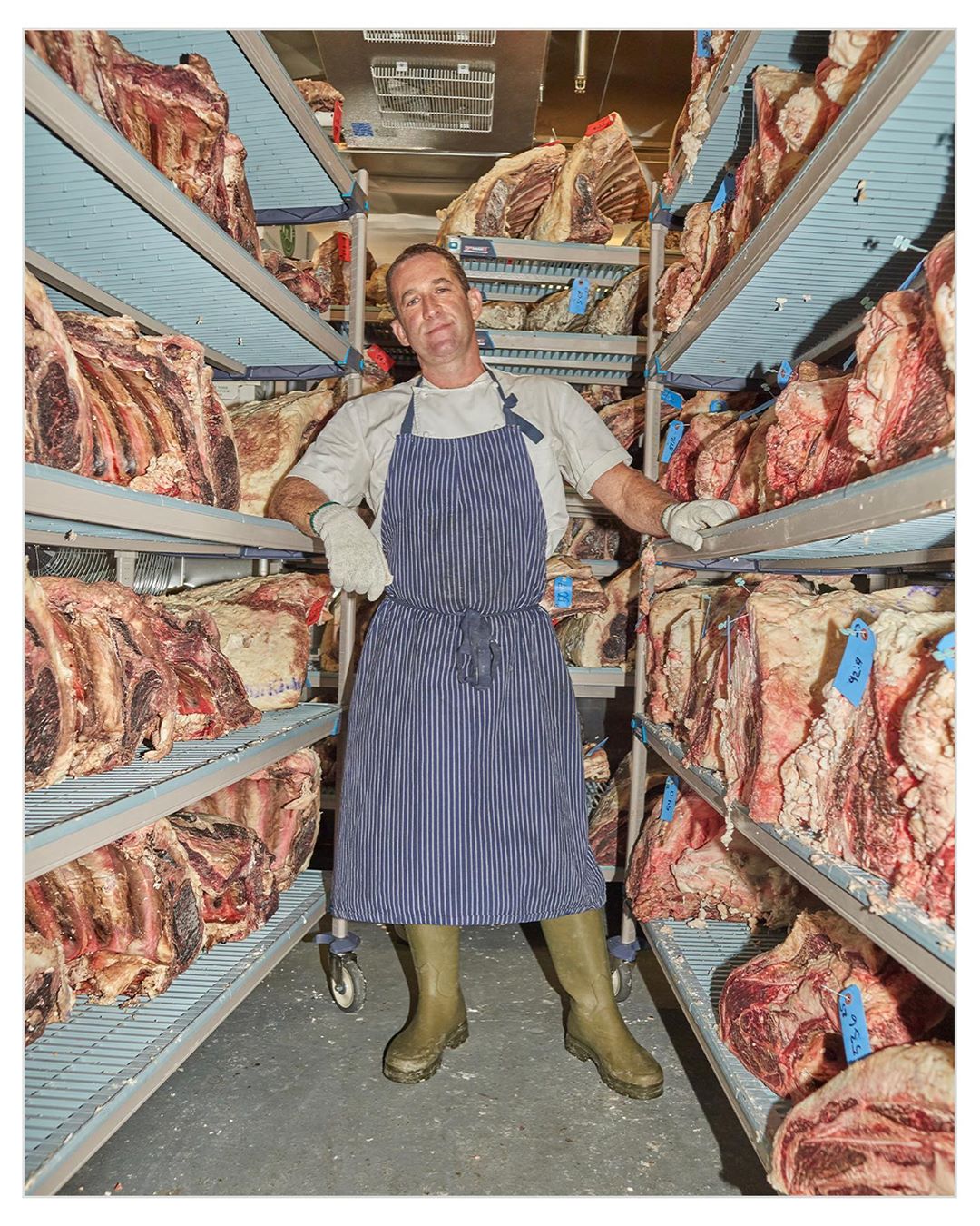 Adult man in an apron stands inside a refrigerated storage area surrounded by shelves filled with large cuts of dry-aged raw beef.