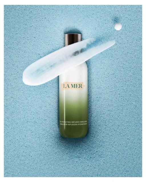 La Mer moisturizer on a blue textured background with a streak of moisturizer and single pearl near the top right corner of the frame.