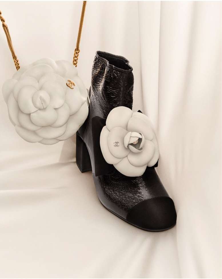 Fashion still life: high-heeled patent leather boot with black toe and white flower appliqué, next to a white flower-shaped purse with gold chain.