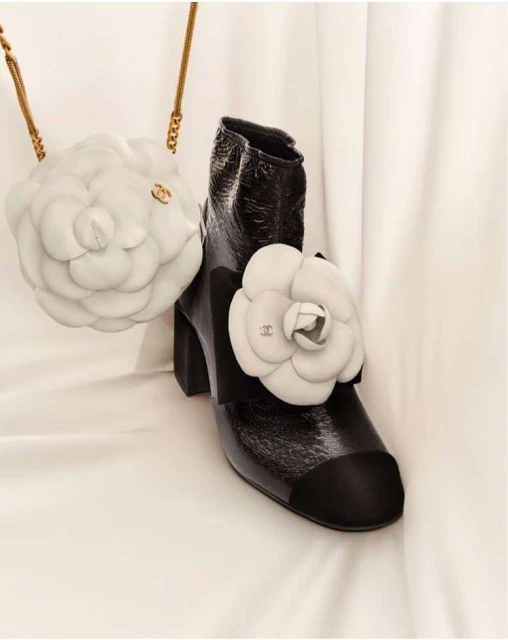 Fashion still life: high-heeled patent leather boot with black toe and white flower appliqué, next to a white flower-shaped purse with gold chain.