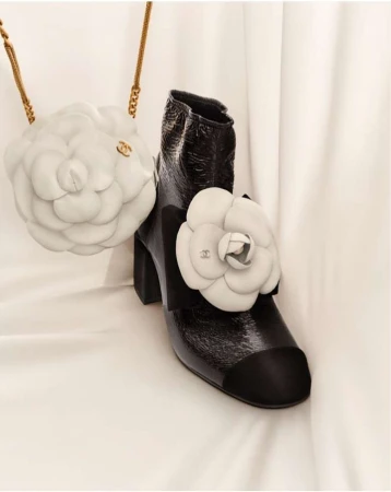 Fashion still life: high-heeled patent leather boot with black toe and white flower appliqué, next to a white flower-shaped purse with gold chain.