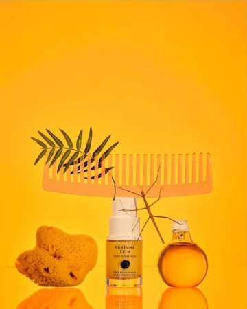 Still life featuring Fortuna Skin product bottle, sea sponge, hair comb, glass bottle, and plant on a yellow background.