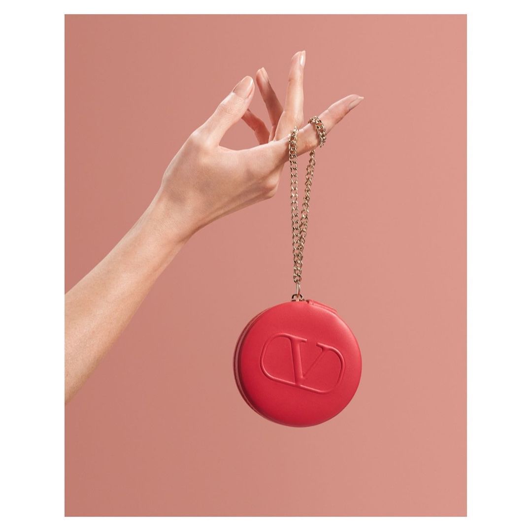 Close-up studio shot of a woman's hand holding a red, round Valentino Garavani clutch on a gold chain against a soft pink background.