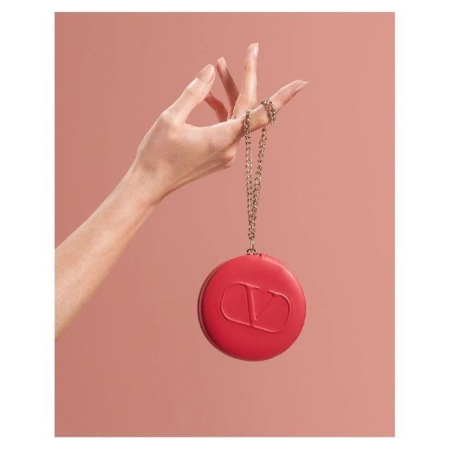 Close-up studio shot of a woman's hand holding a red, round Valentino Garavani clutch on a gold chain against a soft pink background.