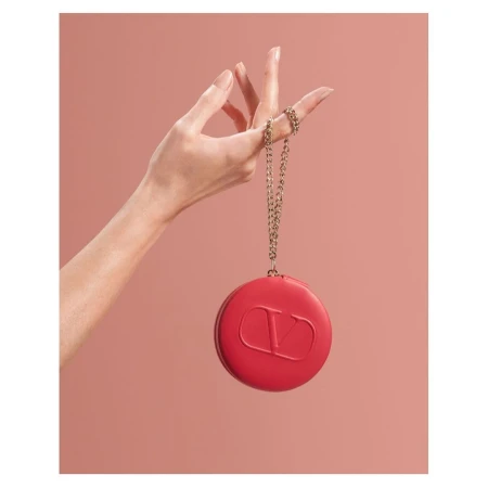Close-up studio shot of a woman's hand holding a red, round Valentino Garavani clutch on a gold chain against a soft pink background.