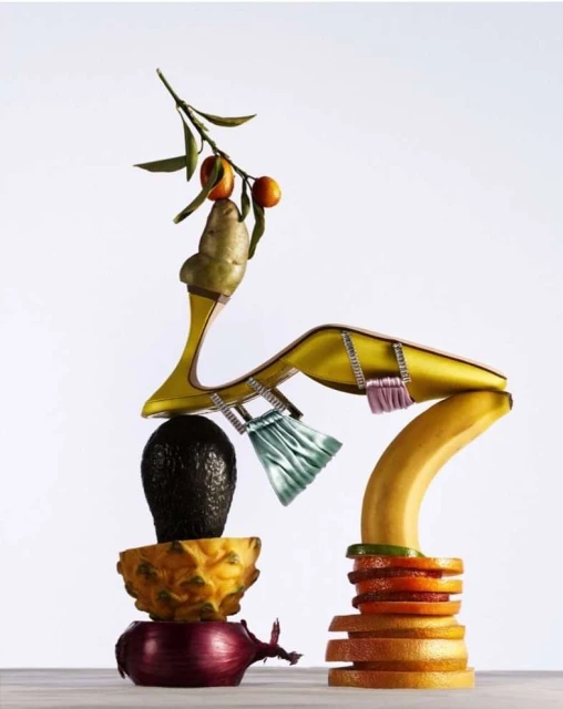 Surreal still life of a yellow high heel shoe balanced on fruits and vegetables, featuring avocados, oranges, and a potato.