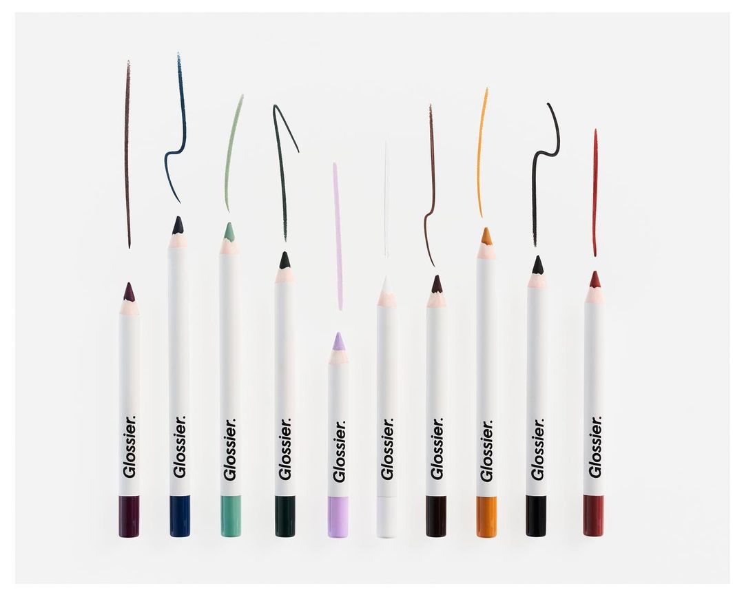 Cosmetic still life featuring a lineup of colorful Glossier eye pencils with matching colored pencil strokes, against a white backdrop.