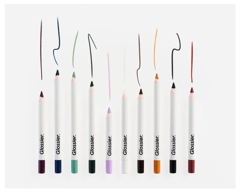 Cosmetic still life featuring a lineup of colorful Glossier eye pencils with matching colored pencil strokes, against a white backdrop.