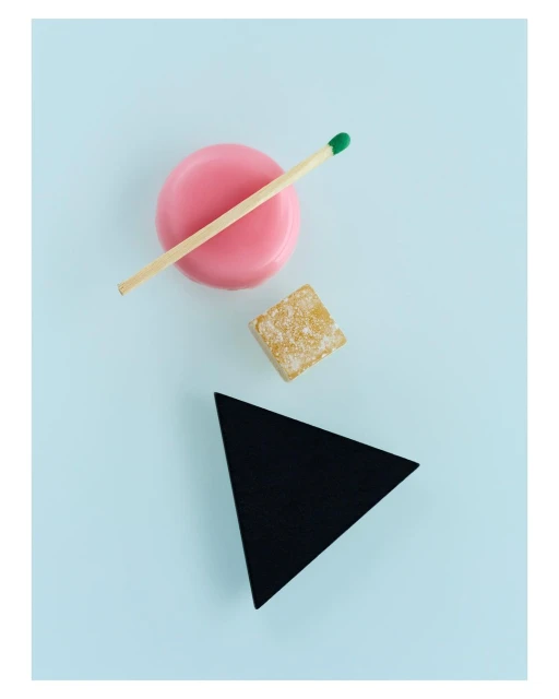 Overhead shot of pink disc, lit matchstick, sugary cube, and black triangle against a light-blue background.