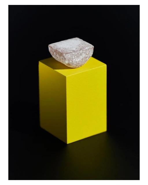 Still life photo of a light brown cube topped with white sugar-like powder on a vibrant yellow cubic pedestal against a stark black background.