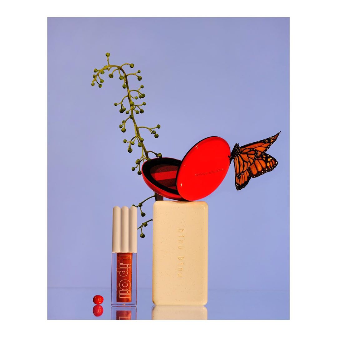 Still life of lip oil, red compact, and cream soap bar balanced with butterfly and floral accents against soft purple background.