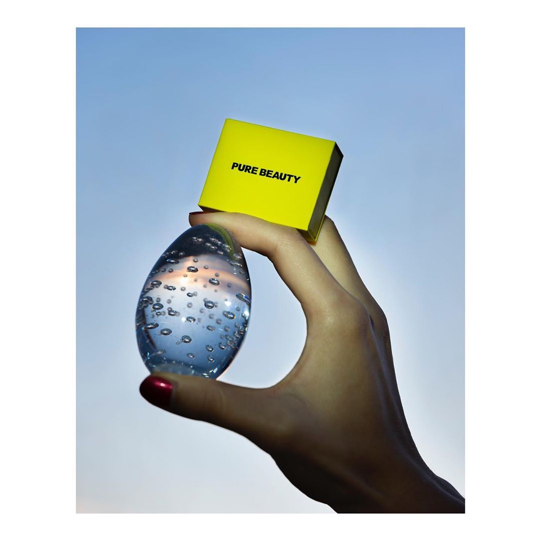 Close-up studio shot of a person's hand holding a clear decorative egg and a yellow beauty product box against a gradient blue background.