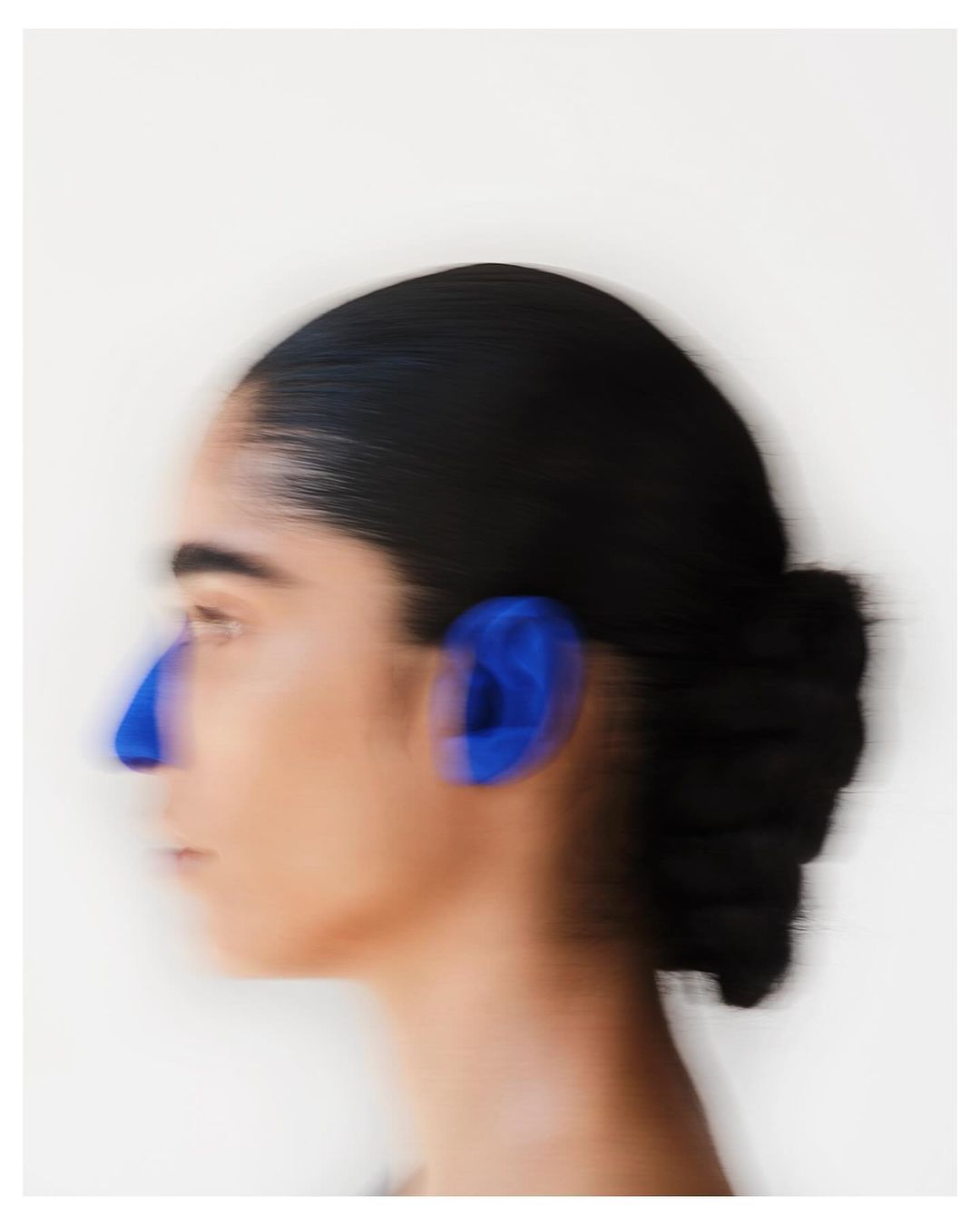 A blurred profile of an adult female with black hair in a bun and blue markings on her face against a plain white background.