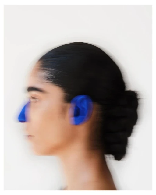 A blurred profile of an adult female with black hair in a bun and blue markings on her face against a plain white background.