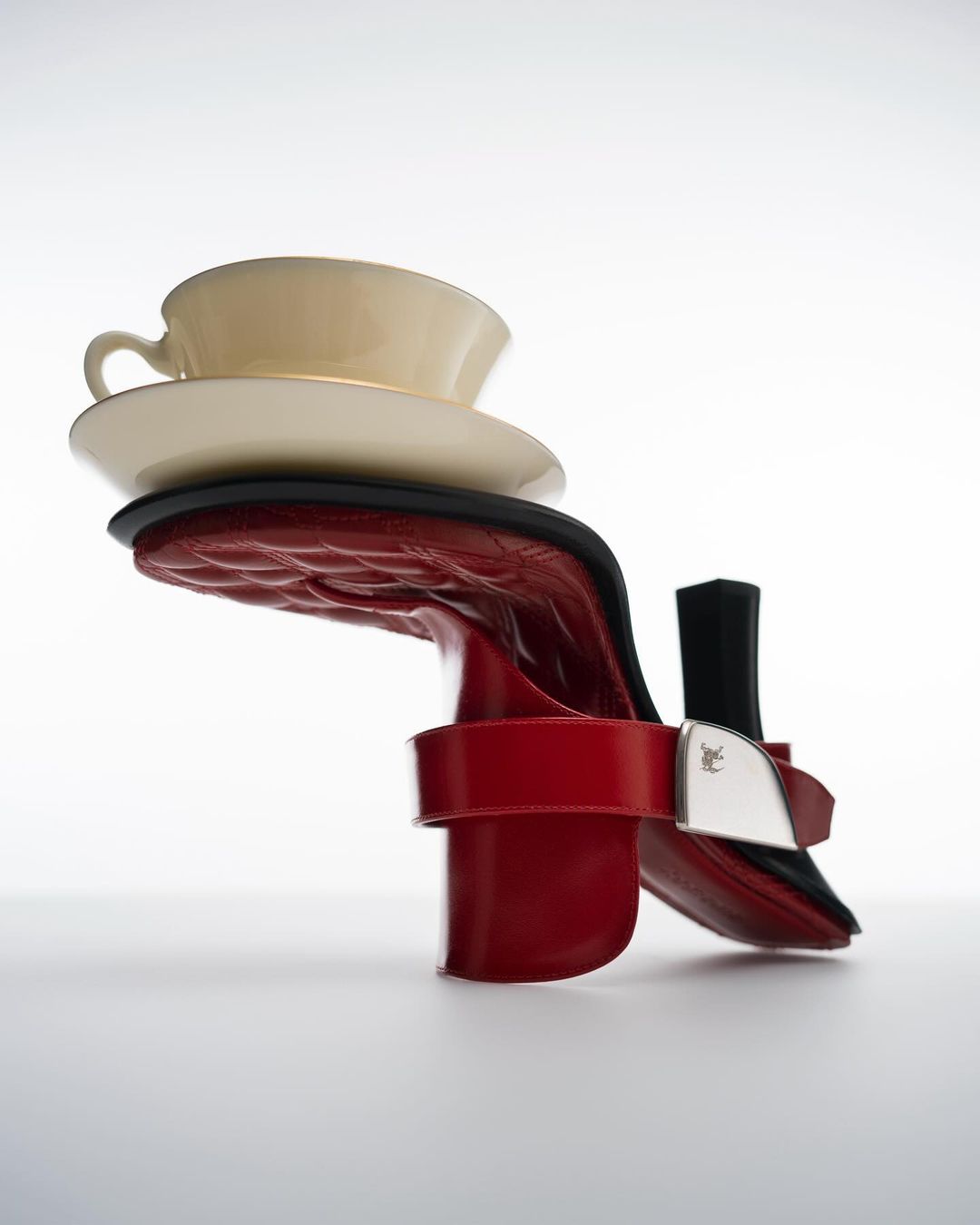 Still life showcasing a red high-heeled shoe balancing a cream-colored teacup and saucer against a clean white backdrop in a studio setting.